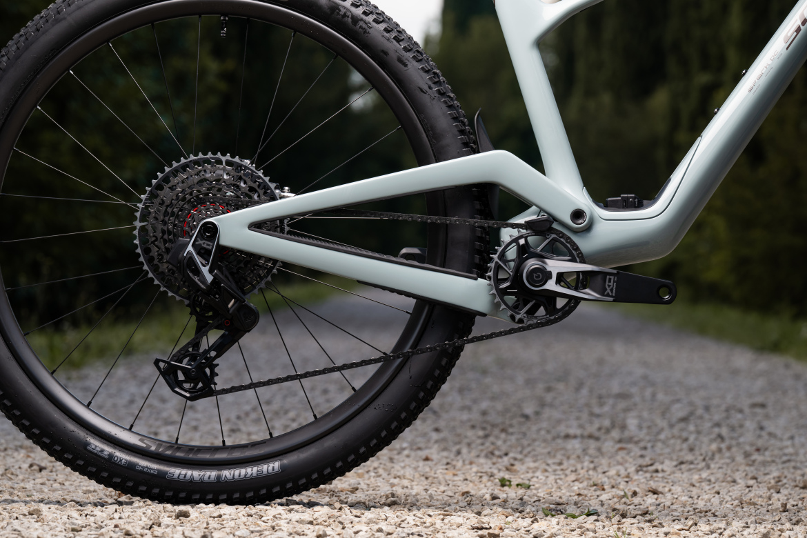 Close-up of a mountain bike's rear drivetrain, showcasing the chain, cassette, and rear derailleur. The bike features a light-colored frame with sleek lines, set against a gravel path and greenery in the background.