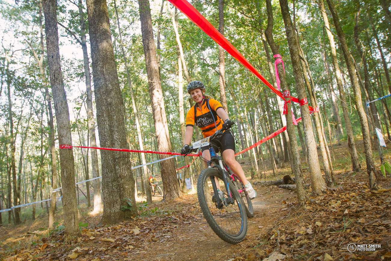 A female mountain biker in an orange and black jersey rides along a dirt trail through a wooded area, navigating between red ribbons marking a course. She has a smile on her face, wearing a helmet, and appears focused and energetic as she rides past tall trees and fallen leaves on the ground.