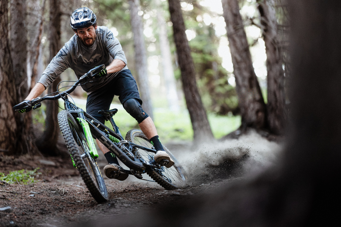 A mountain biker skillfully navigates a dirt trail in a forest, leaning into a turn while kicking up dust. The rider is wearing a helmet and protective gear, and the scene is surrounded by tall trees, emphasizing the natural outdoor setting.