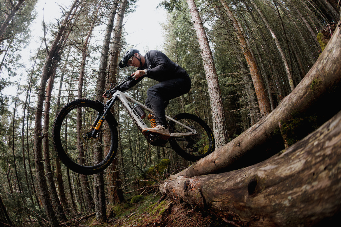 A mountain biker performing a jump over a fallen log in a dense forest, surrounded by tall pine trees. The rider is wearing a helmet and athletic gear, showcasing a dynamic pose as they navigate the rugged terrain.