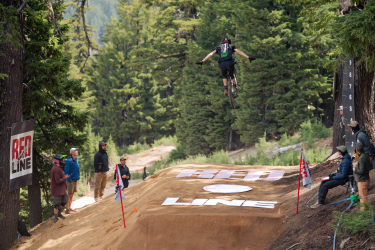 A mountain biker in mid-air over a dirt jump, surrounded by a forested landscape. Spectators watch from the side, with banners displaying the "Red Line" logo visible in the foreground. The scene captures the excitement of biking sports in a natural setting.