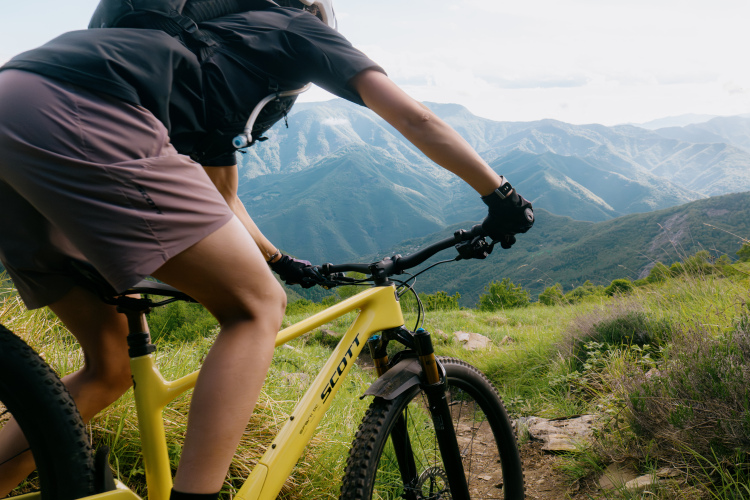 A cyclist riding a yellow mountain bike on a grassy trail, leaning forward to navigate the terrain. The background features a panoramic view of green mountains under a partly cloudy sky. The cyclist is wearing a helmet and a black shirt, with a hydration pack on their back.