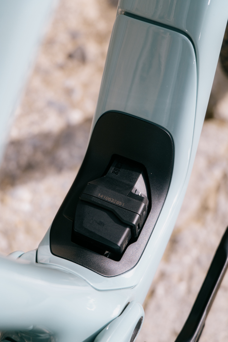 Close-up of a bike's frame showing a secured battery compartment with a black connector nestled in a matte black surface against a light blue backdrop. The background is blurred, revealing a natural setting.