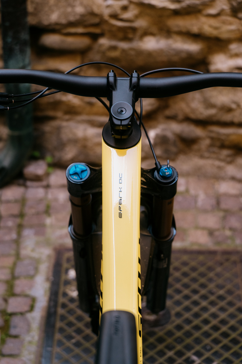 Close-up view of a mountain bike, focusing on the handlebars and front fork. The frame is bright yellow with the name "SPARK" displayed prominently. The handlebars are black with visible brake and gear cables. The background features a textured stone wall and a patterned surface below the bike.