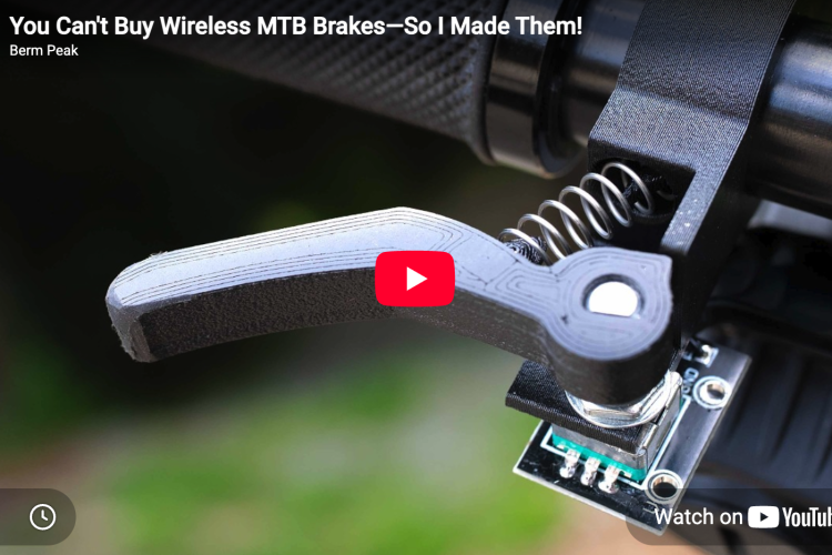 Close-up image of a mountain bike brake lever and its mechanical components, including a spring and electronic circuit board. The background is blurred greenery, and a play button indicates this is a still from a YouTube video titled "You Can't Buy Wireless MTB Brakes&mdash;So I Made Them!" from the channel Berm Peak.