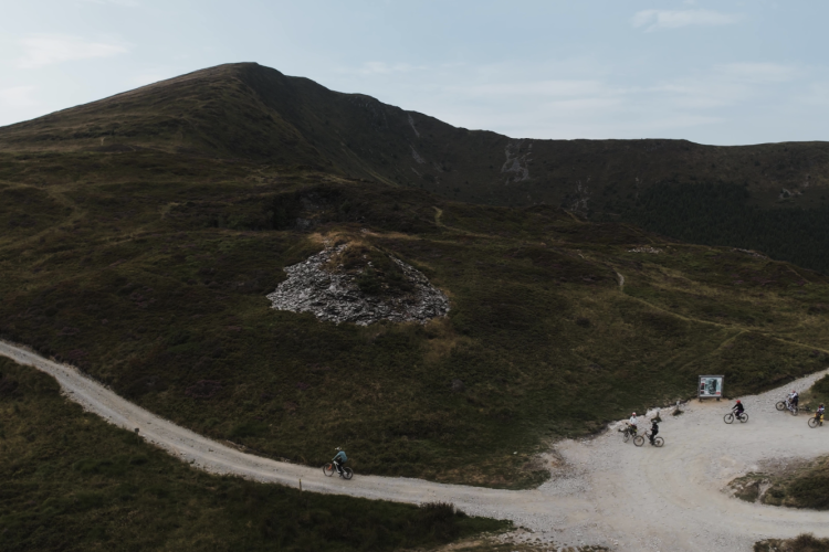 Aerial view of a mountainous landscape featuring dirt bike trails and several cyclists navigating the terrain. Green hills are dotted with rocky outcrops under a clear sky.