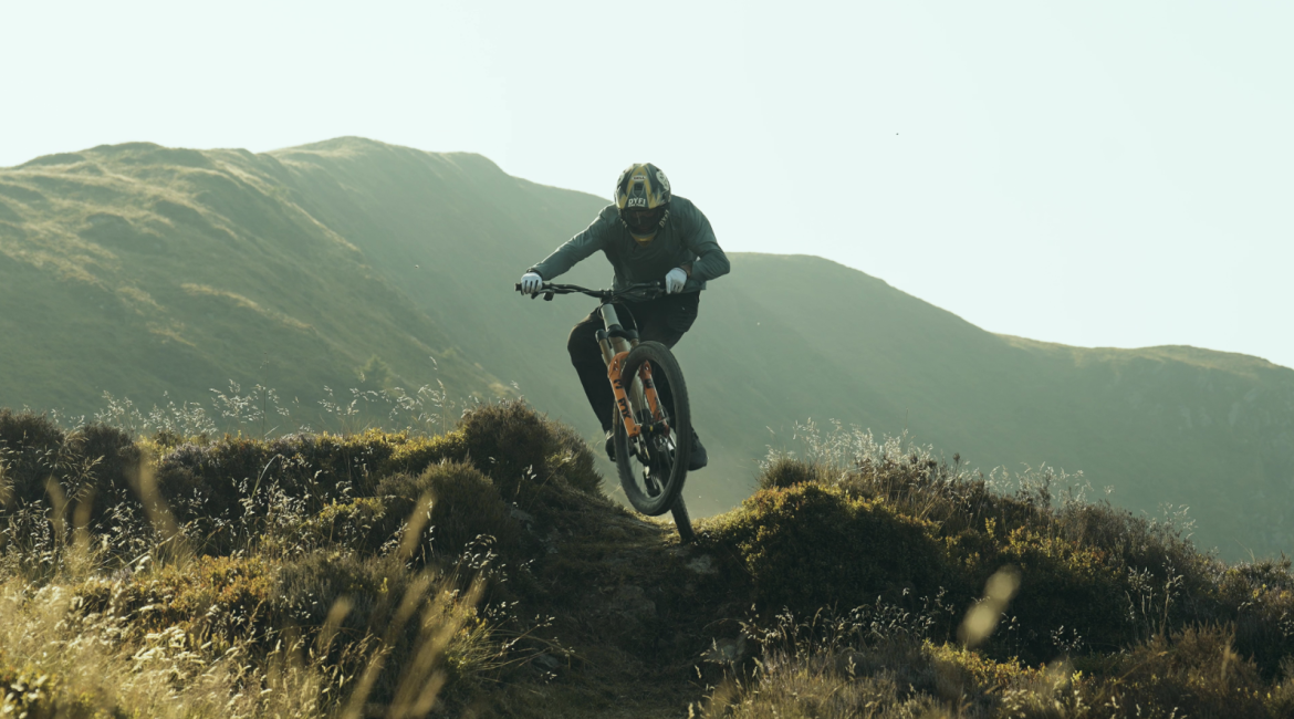 A mountain biker in protective gear performs a jump on a rocky trail, surrounded by rolling hills and grassland in the background. The scene captures the energy and excitement of outdoor biking in a natural landscape during daylight.