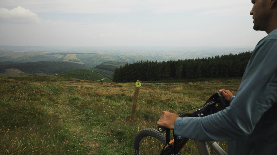 A person standing on a grassy hillside, holding a mountain bike and looking out over a scenic landscape of rolling hills and forests under a cloudy sky. A trail marker is visible in the foreground, indicating a biking path.