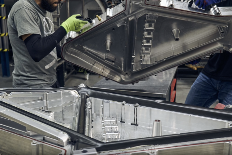 Two workers wearing gloves are handling a large, detailed aluminum casing in a manufacturing environment. The casing is open, revealing intricate interior components and designs. The setting appears to be a workshop or factory, with various tools and machinery in the background.