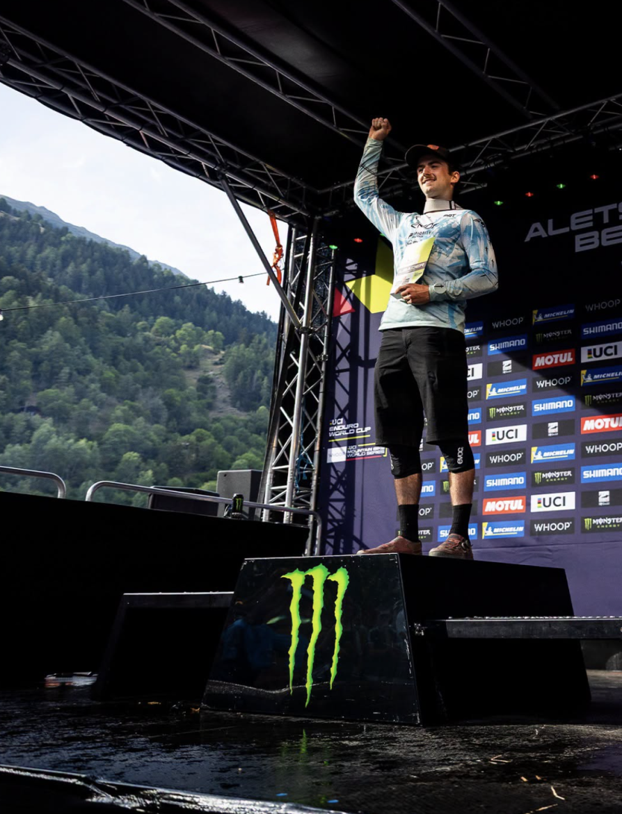 A cyclist stands on a podium with one fist raised in celebration, wearing a light blue shirt and black shorts. The backdrop features logos of sponsors, and the scenery includes green hills in the background. The platform has a prominent Monster Energy logo.