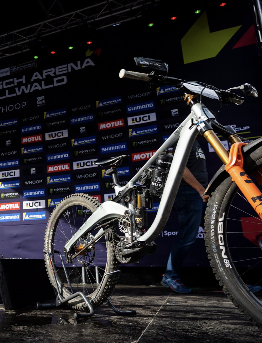 A white mountain bike with prominent suspension and thick tires stands on a bike stand, positioned in front of a backdrop featuring logos for various sponsors at a cycling event. The background includes colorful lighting and branding elements associated with the UCI Mountain Bike World Cup.