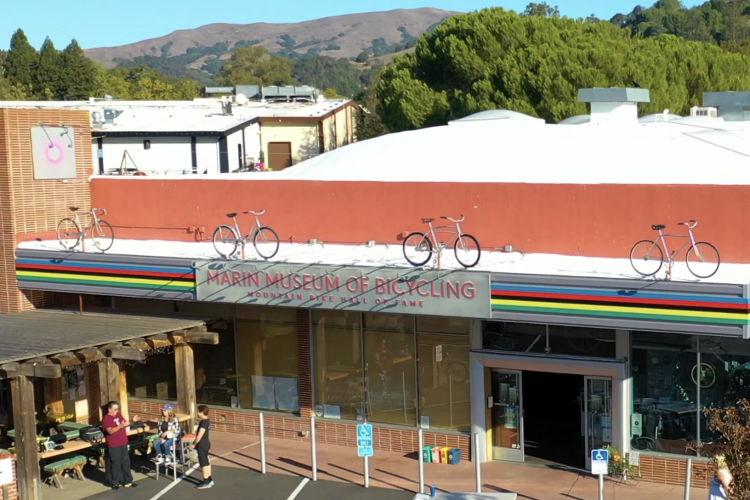 Exterior view of the Marin Museum of Bicycling, featuring a colorful striped awning with bicycles displayed on the roof. In the foreground, a wooden seating area is visible with people gathered, while the backdrop showcases rolling hills and trees. The museum's name is prominently displayed on the building's facade.