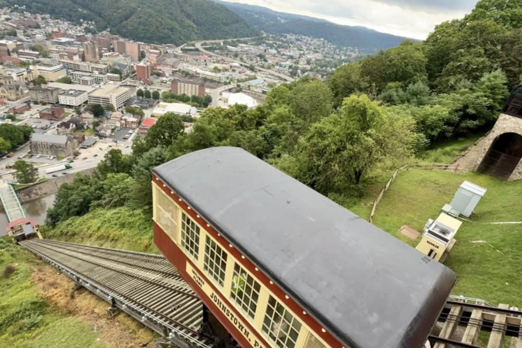 A view of a hillside with a funicular railway descending towards a city below. The train car is positioned on the tracks, surrounded by greenery, with buildings and a river visible in the valley. The landscape features rolling hills and structures amidst a cloudy sky.
