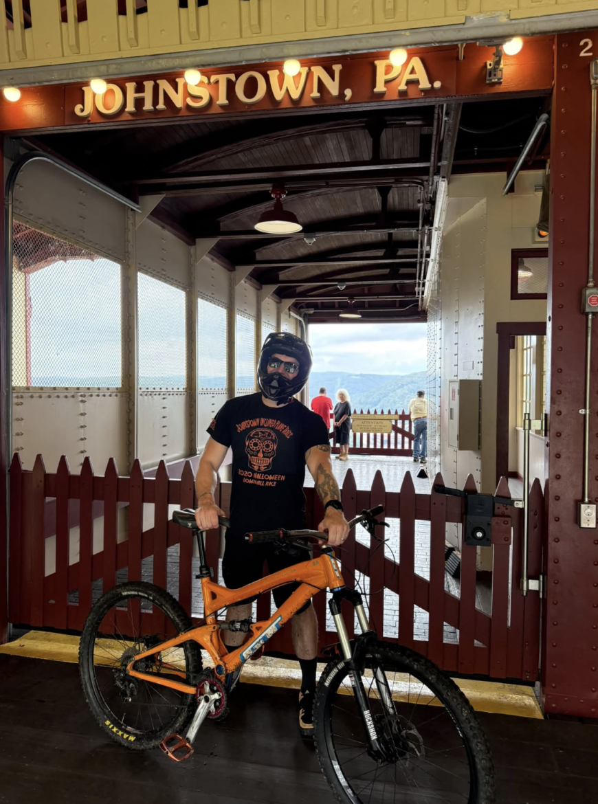 A person wearing a helmet and sunglasses stands with an orange mountain bike in front of the Johnstown, PA sign. The background features a wooden structure with a railing and a few people walking in the distance. The scene captures a mix of recreational biking and a local landmark.