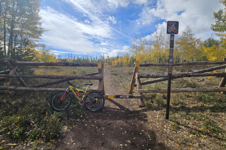 A mountain bike resting against a wooden fence gate in a scenic outdoor area. The background features trees with autumn-colored leaves and a clear blue sky with scattered clouds. A sign on the fence indicates that electric bikes are not allowed on the trail.