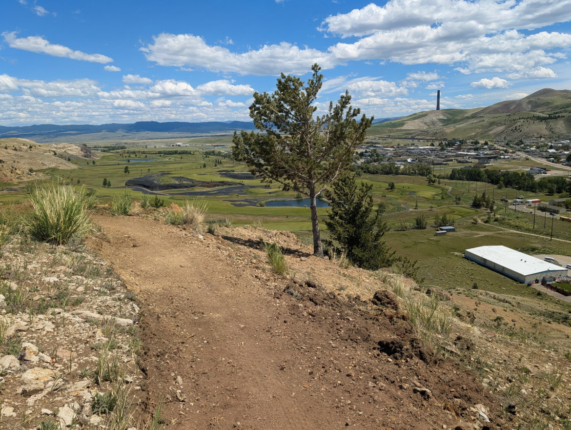 A scenic view from a hillside featuring a dirt path, greenery, and a distant town. The landscape includes fields, patches of water, and mountains under a blue sky with scattered clouds. A prominent industrial structure is visible in the background, adding depth to the rural setting.