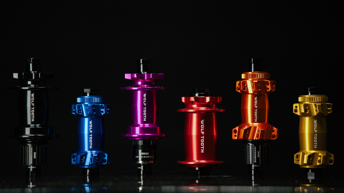 A group of colorful bicycle hubs arranged in a line, featuring black, blue, purple, red, orange, and gold finishes, all labeled with "WOLF TOOTH." The hubs are displayed against a dark background, highlighting their shiny metallic surfaces.