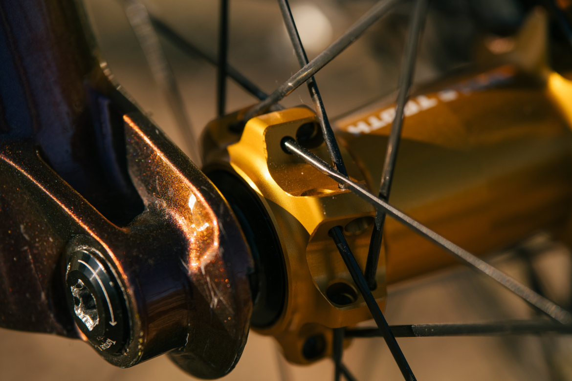 Close-up image of a bicycle wheel hub, featuring a shiny golden hub connected to black spokes, with part of the bike frame visible. The surface reflects light, showcasing metallic textures and intricate details of the components.