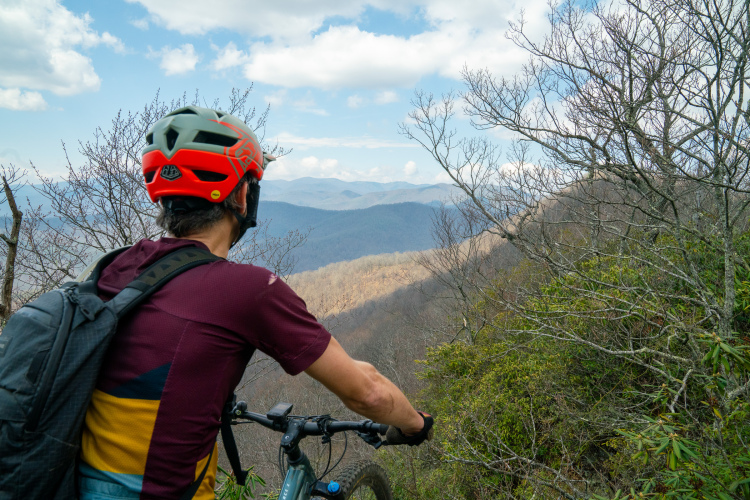 A person wearing a red and grey bicycle helmet and a maroon cycling jersey is looking out over a mountainous landscape. The scene features rolling hills with sparse trees, under a sky filled with scattered clouds. The individual is holding onto their bicycle while taking in the view.