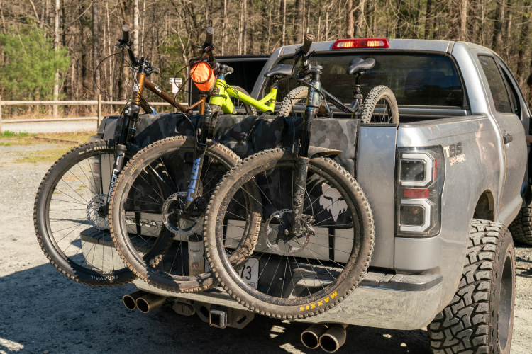 Alt text: Two mountain bikes secured in the bed of a pickup truck, positioned against a backdrop of trees. The bikes feature various colors and designs, including a bright yellow frame, with dirt on their tires indicating use. The truck is parked on a gravel surface, suggesting a location suitable for outdoor activities.