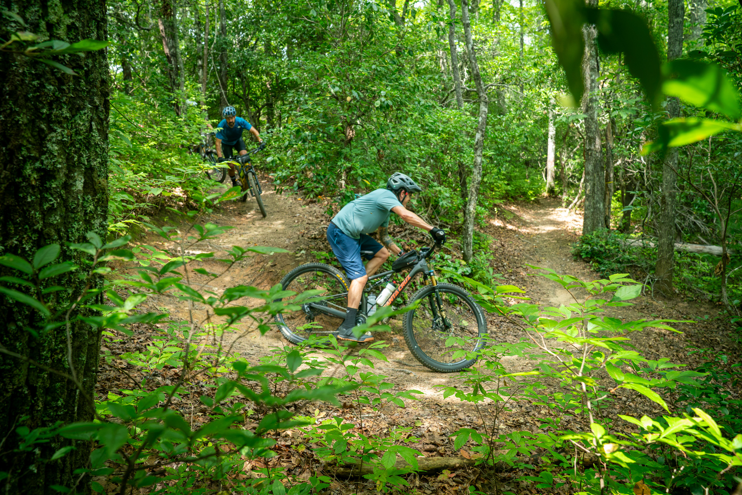 Two mountain bikers riding on a dirt trail surrounded by lush greenery and trees. The first rider, in a blue shirt and helmet, is navigating a curve, while the second biker can be seen in the background. The scene captures the essence of outdoor adventure and mountain biking in a forested area.