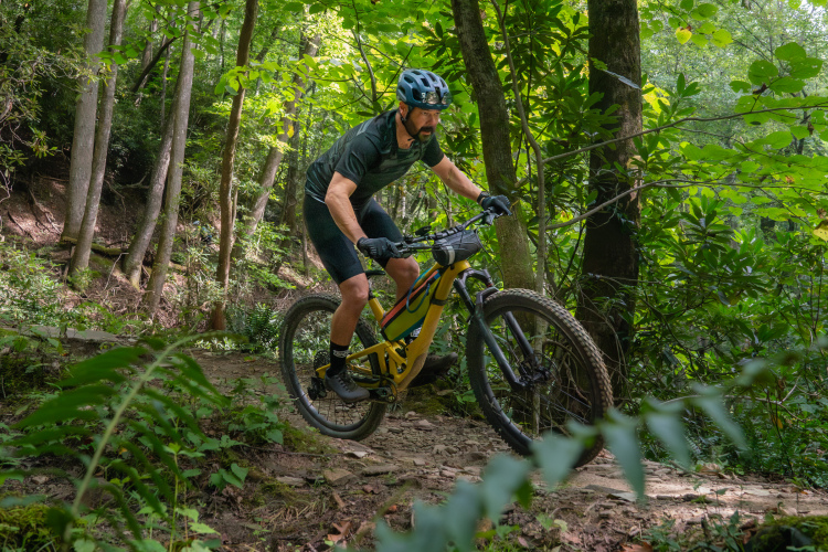 A mountain biker rides a yellow bike along a narrow trail surrounded by lush green foliage and trees. The cyclist is focused and wearing a helmet, navigating the rocky terrain in a wooded area.