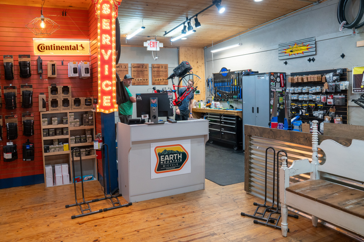 Alt text: An interior view of a bicycle shop featuring a service counter with a sign that says "SERVICE." The shop displays various bicycle accessories on the walls, including tires and tools, with a wooden floor and warm lighting. Two employees are engaged in bicycle maintenance in the background.