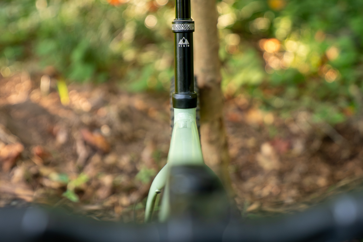 Close-up view of a bicycle's seat post and frame, with a blurred background of greenery and fallen leaves. The seat post features a black design with a mountain logo, set against a soft-focus natural environment.