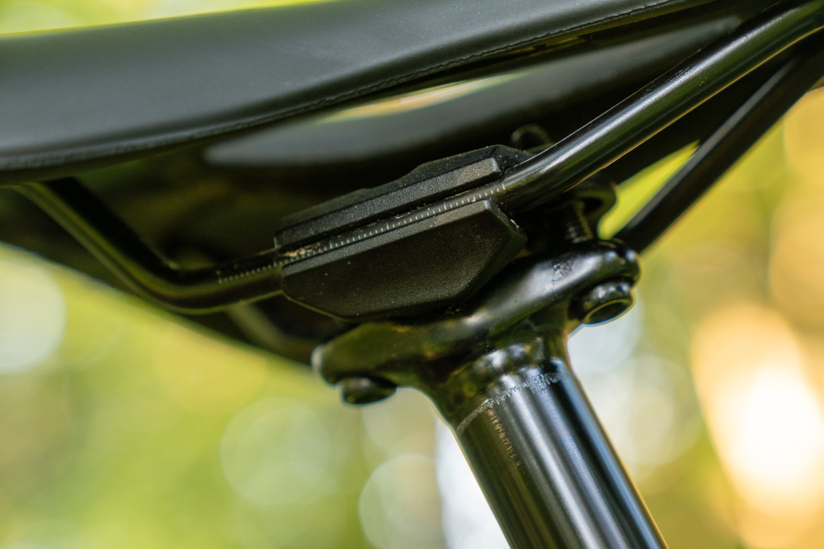 Close-up image of a bicycle saddle and seat post, showcasing the saddle attachment mechanism. The background features a soft, blurred green bokeh effect, indicating an outdoor setting.