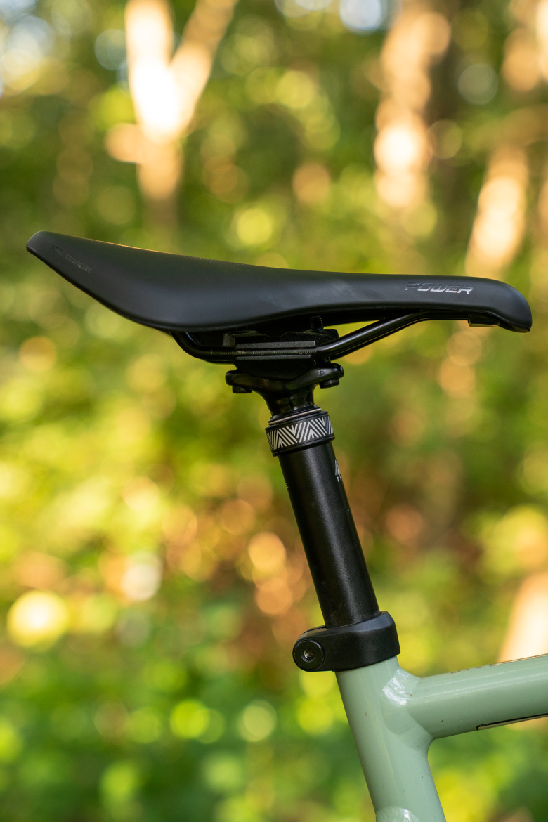 A close-up of a black bicycle saddle mounted on a black seat post, with a decorative collar, against a blurred green and brown background of trees and foliage.