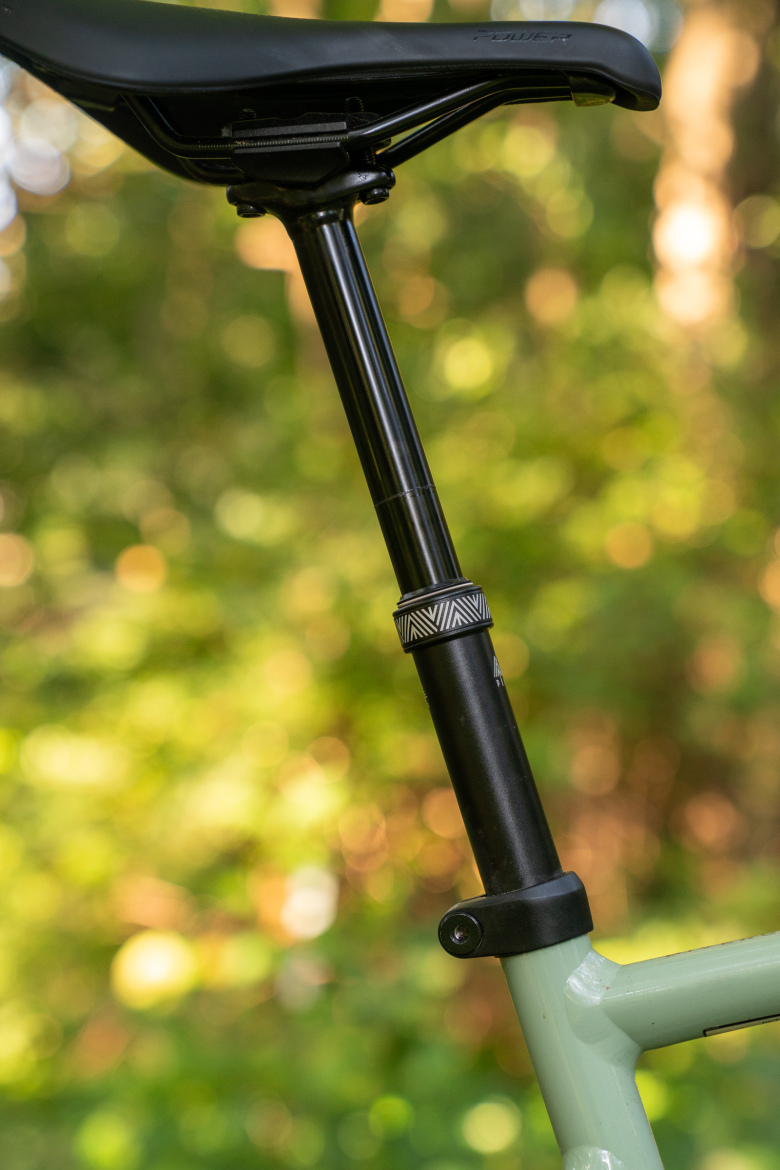 Close-up of a bicycle seat post and saddle against a blurred green background. The seat post is black with a patterned adjustment collar, connected to a light green bike frame. The image captures the details of the components in a natural outdoor setting.