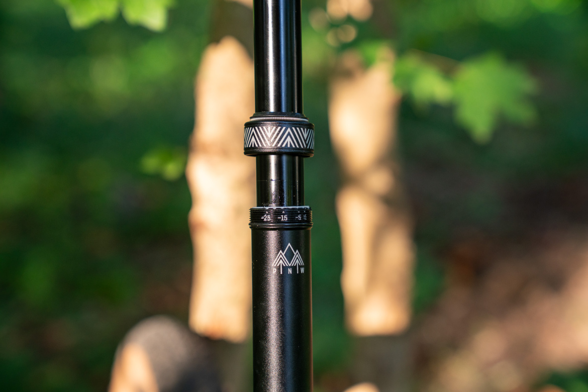 Close-up of a black adjustable bicycle seat post with measurement markings and a geometric patterned adjustment collar. The background features soft focus greenery, creating a natural outdoor setting.