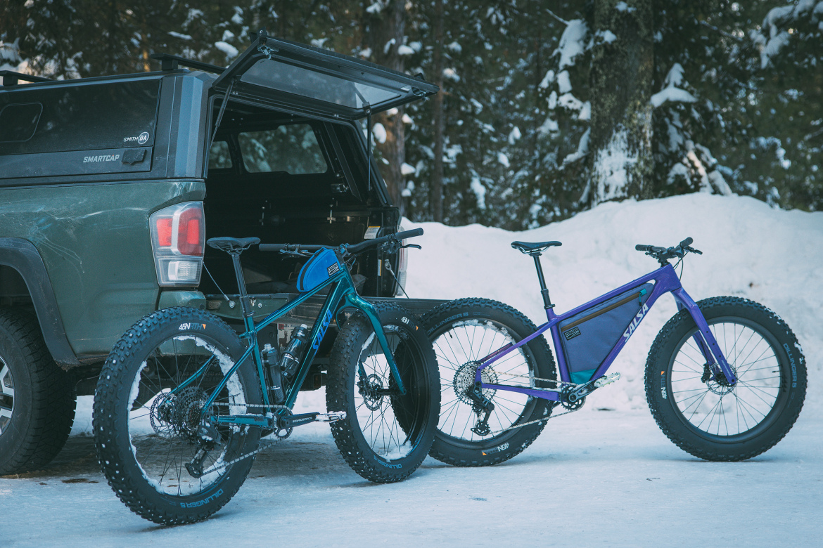 A green pickup truck with its rear door open, showcasing two fat tire bicycles parked on a snowy surface. The bicycles include a teal bike with mountain bike features and a purple bike with a distinctive frame design. Pine trees are visible in the background, creating a wintery setting.