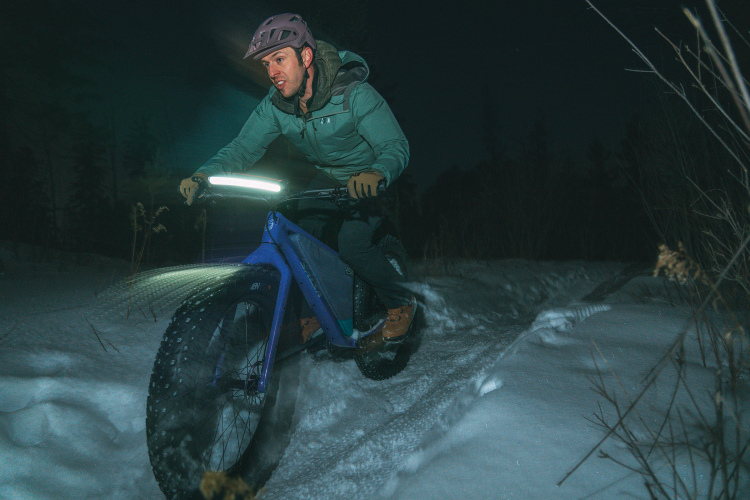 A man rides a fat tire bike on a snowy trail at night, wearing a helmet and a jacket. The bike has a light attached, illuminating the path ahead. Trees are visible in the background, creating a serene winter landscape.
