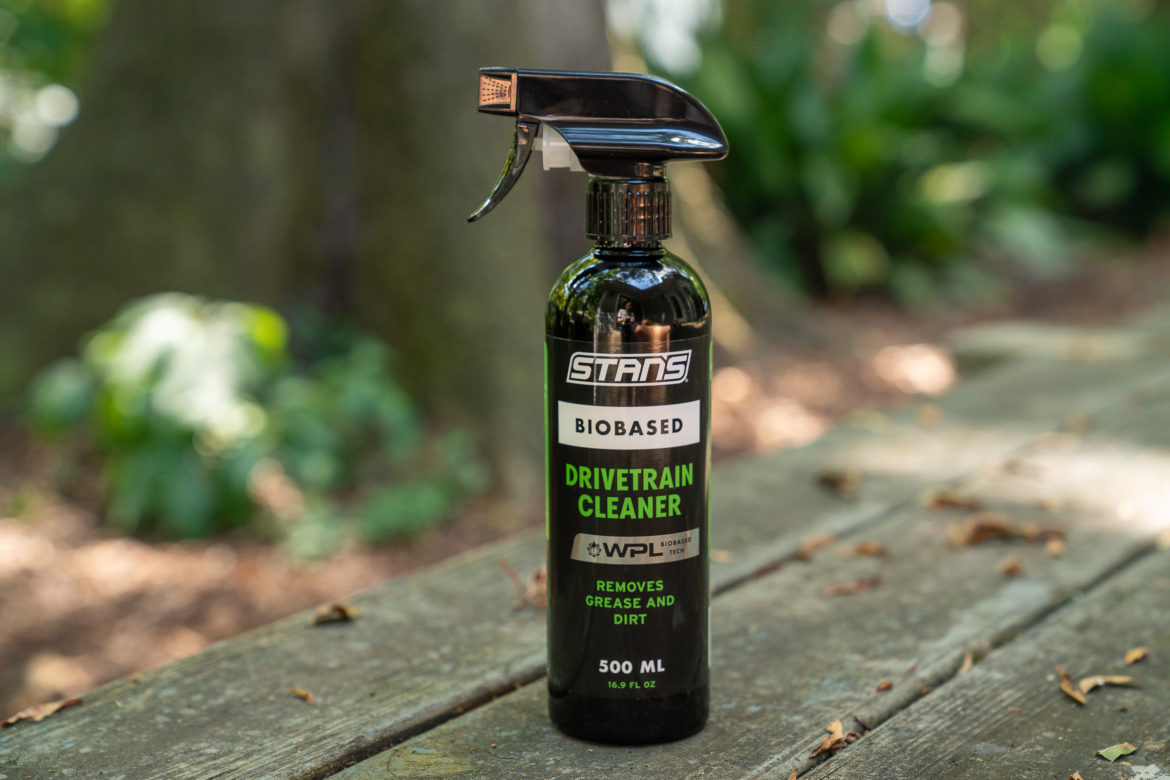 A bottle of Stan's Biobased Drivetrain Cleaner, 500 mL, with a black spray nozzle, displayed on a weathered wooden surface surrounded by greenery. The label highlights its ability to remove grease and dirt.