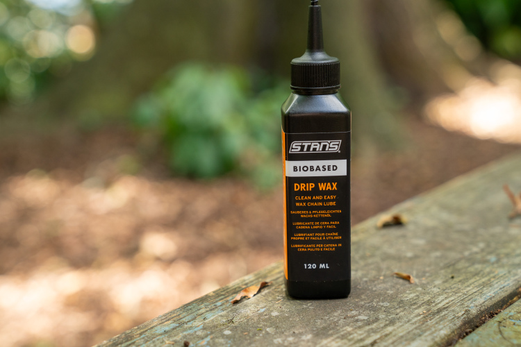 A bottle of STANS Biobased Drip Wax chain lube sitting on a wooden surface. The label highlights its features, including "clean and easy wax chain lube," and indicates the product volume as 120 mL. The background is blurred, featuring greenery and natural elements.