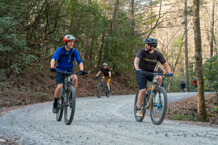Two mountain bikers smile and converse on a gravel path in a wooded area, while others ride in the background. The scene captures a sense of camaraderie and outdoor adventure amidst trees and natural surroundings.