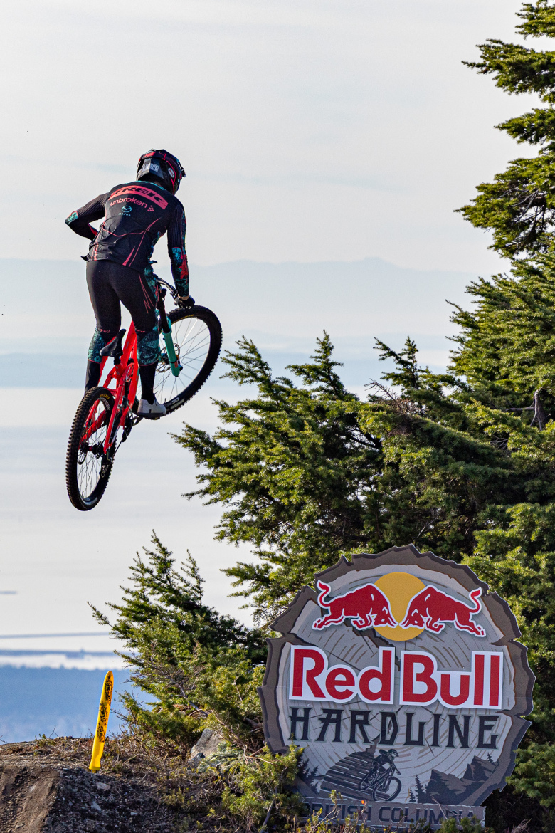 A mountain biker in a black and colorful outfit jumps off a rocky ledge, with a scenic view of the landscape in the background. The biker is airborne in front of a large sign that reads "Red Bull Hardline," surrounded by evergreen trees.
