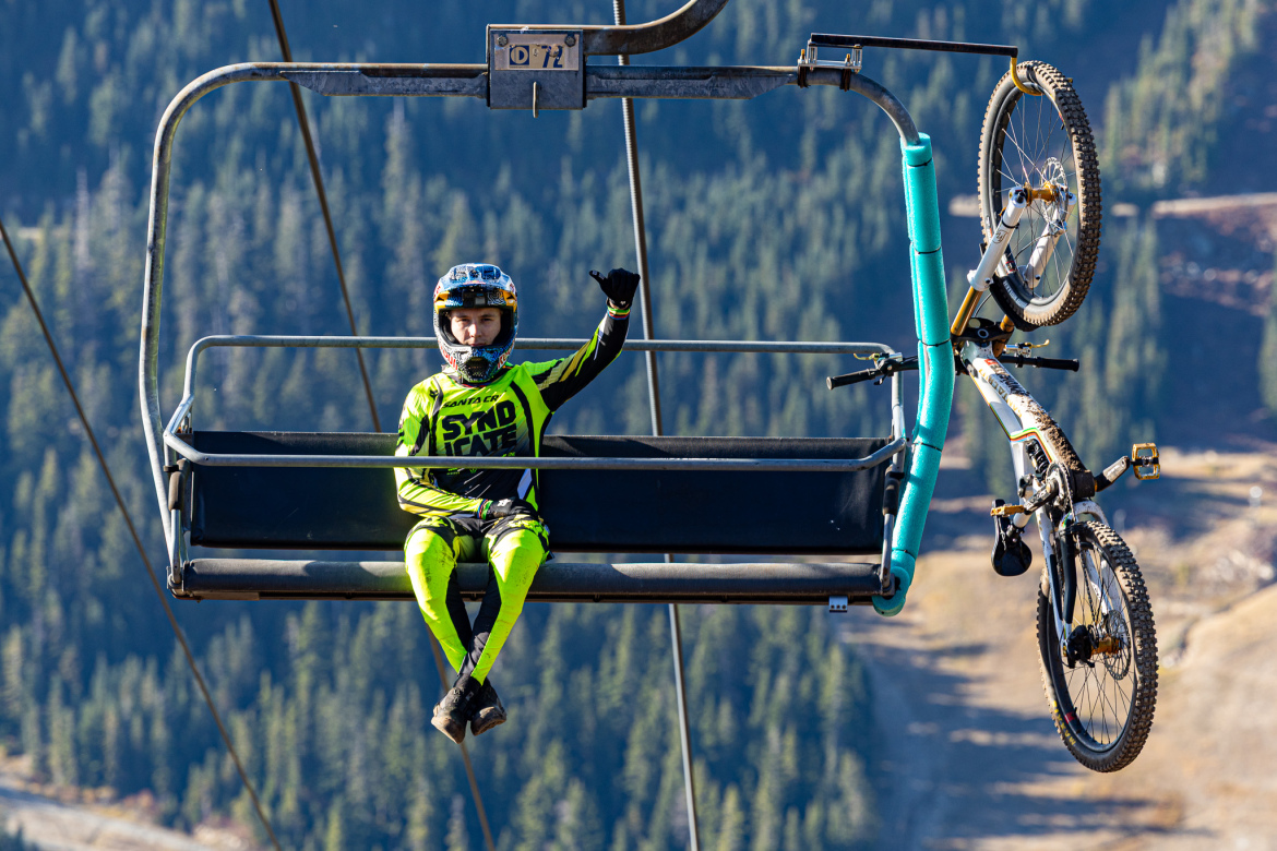 A young cyclist in a bright yellow and black outfit sits on a ski lift chair with a mountain landscape in the background. The cyclist, wearing a helmet, is giving a thumbs up with one hand while the other rests on his knee. A mountain bike is attached to the lift next to him, its front wheel hanging down. The scene captures a moment of excitement and adventure in a mountainous outdoor setting.