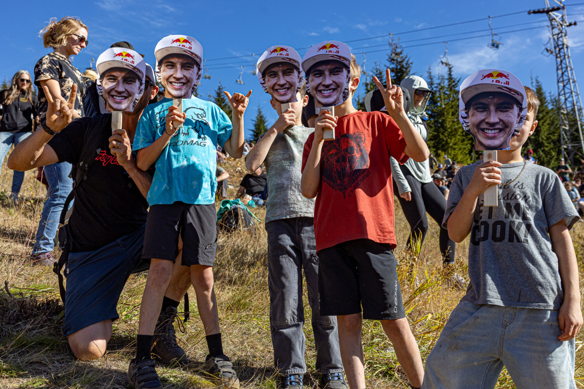 A group of children and an adult pose happily outdoors, wearing large cut-out masks of a smiling face and holding signs. They are on a hillside with spectators in the background, enjoying a sunny day with trees and power lines visible. The adult kneels in front, making a hand gesture, while the children show excitement with peace signs and cheerful expressions.