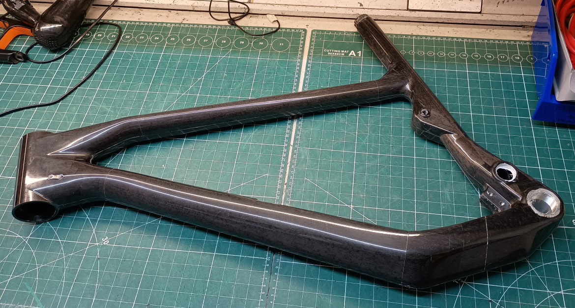 A carbon fiber bicycle frame positioned on a green cutting mat, with various tools and equipment in the background. The frame features a sleek design and is partially visible, highlighting its intricate structure and finish.