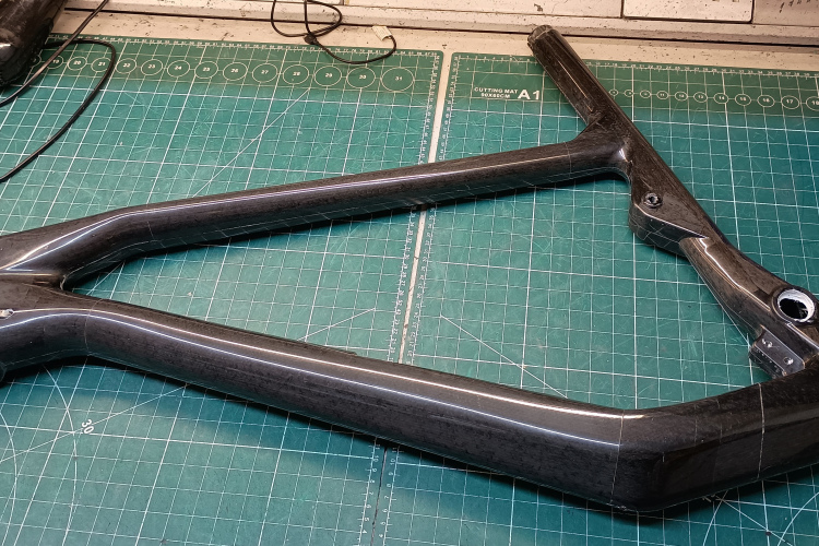 A carbon fiber bicycle frame positioned on a green cutting mat, with various tools and equipment in the background. The frame features a sleek design and is partially visible, highlighting its intricate structure and finish.