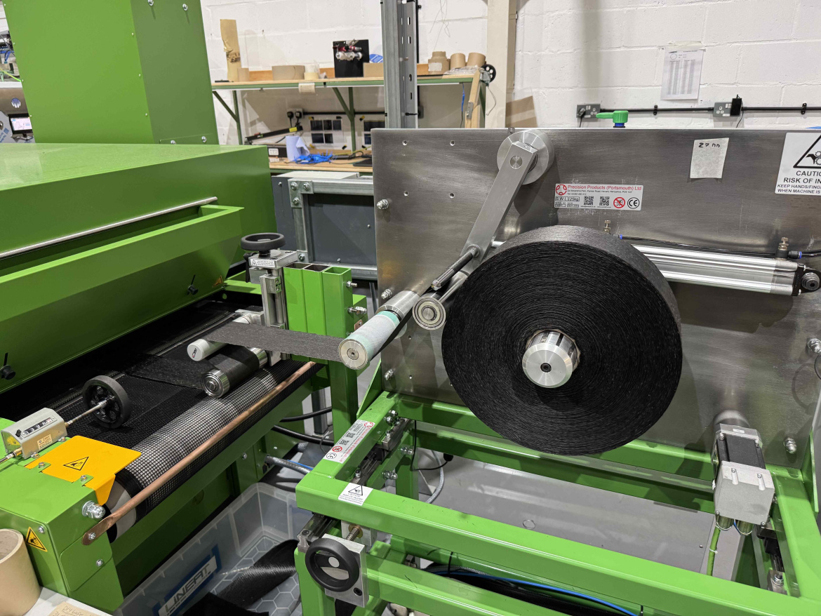 Industrial machinery featuring a green and silver design, with a roller mechanism and a black spool. The setup includes a conveyor belt and various components for processing materials, situated in a factory or workshop environment.
