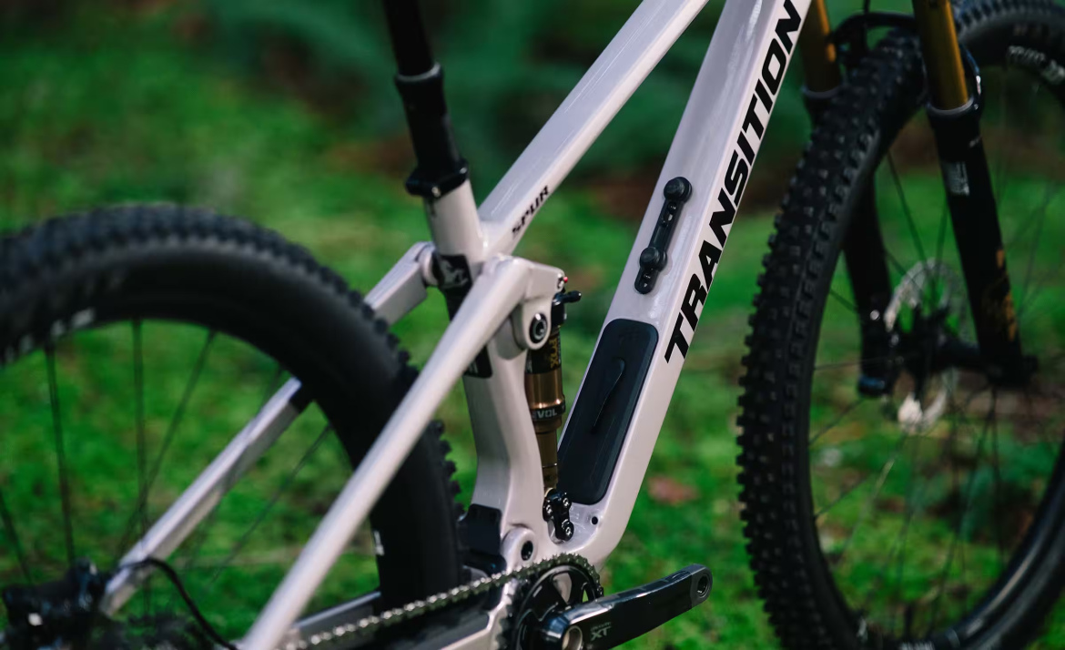 Close-up of a Transition mountain bike frame in a natural setting, showcasing its sleek design, including the suspension components and drivetrain details, against a backdrop of green grass.