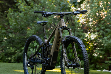 A mountain bike positioned on green grass, surrounded by lush foliage. The bike features a sleek design with a dark frame and prominent tires, showcasing its suitability for off-road biking.