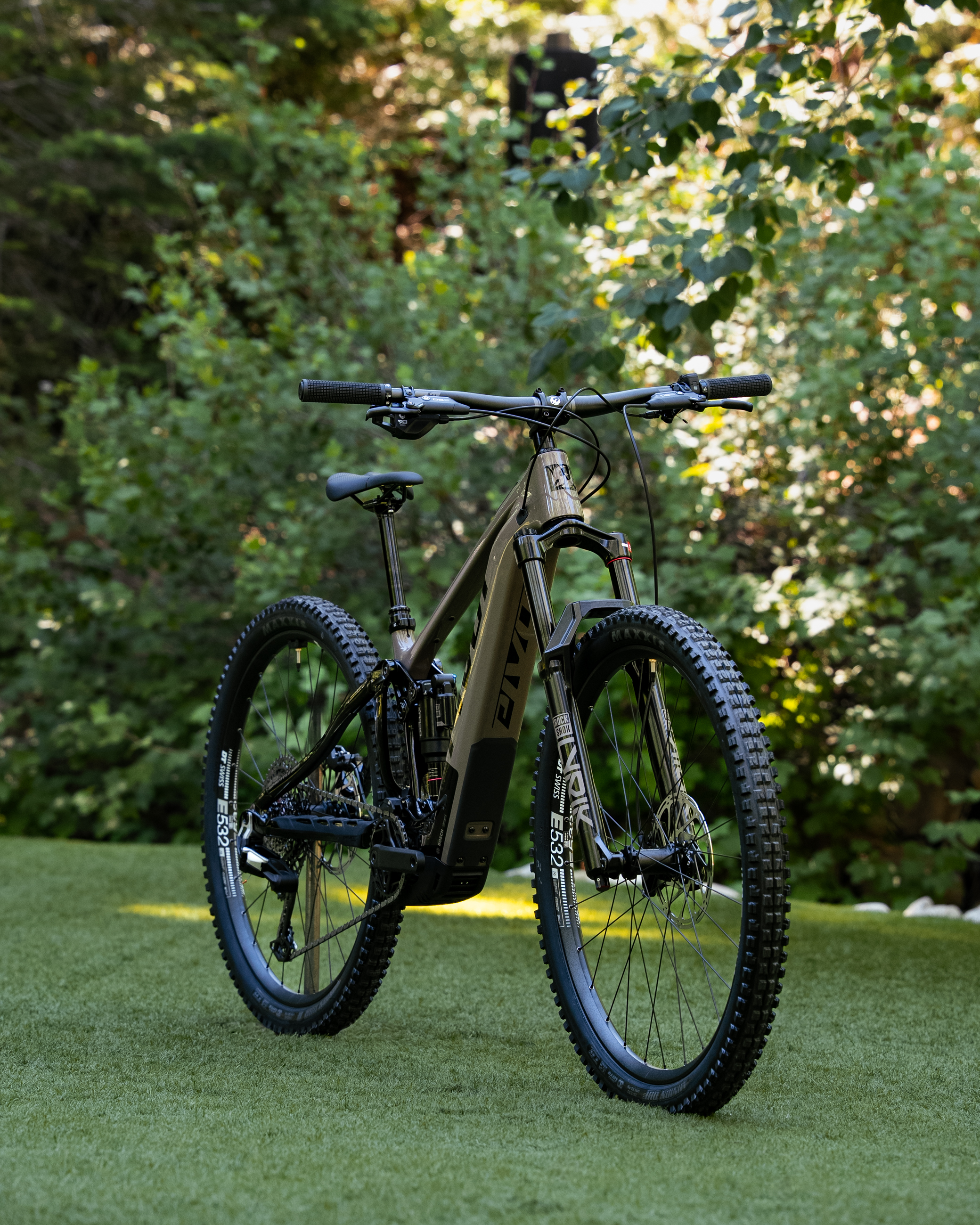 A mountain bike positioned on green grass, surrounded by lush foliage. The bike features a sleek design with a dark frame and prominent tires, showcasing its suitability for off-road biking.