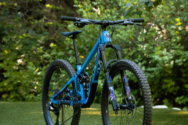 A blue mountain bike with thick tires and suspension forks, positioned on a green grassy surface with lush foliage in the background.
