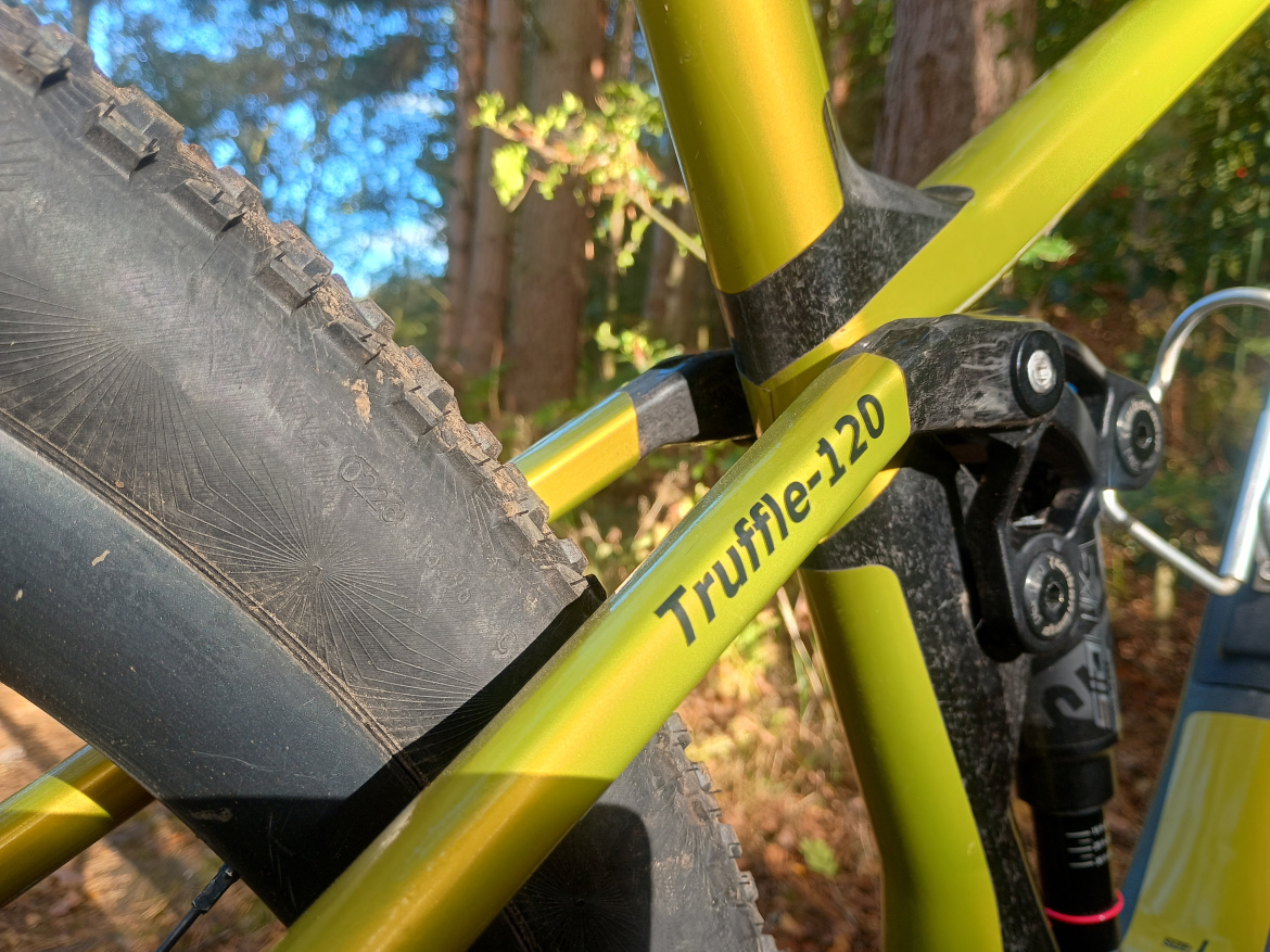 Close-up image of a mountain bike frame with the name "Truffle-120" visible, showcasing a yellow and black color scheme, with a focus on the rear tire and suspension components. The bike is positioned in a wooded area, with soft natural light filtering through the trees.