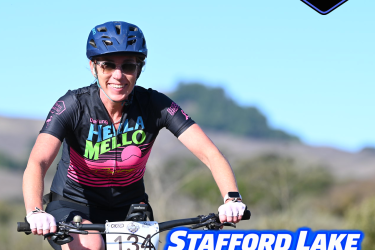 A smiling cyclist wearing sunglasses and a colorful jersey rides a mountain bike on a sunny day. The image includes text that reads "November 8, 2026," "Stafford Lake Rattler XC," and "+ Juniors Fundraiser," along with a logo in the corner. The background features a scenic view of hills and greenery.