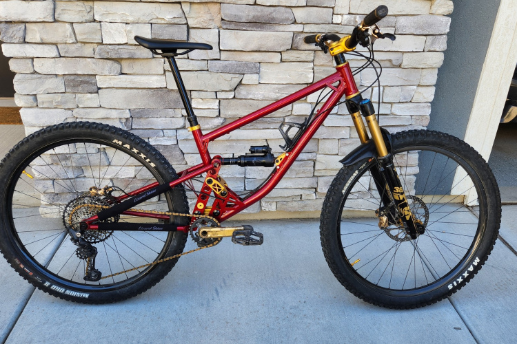 Mountain bike with a red frame, featuring gold accents, thick tires, and a front suspension fork. The bike is parked on a concrete surface against a stone wall, showcasing its design and components.